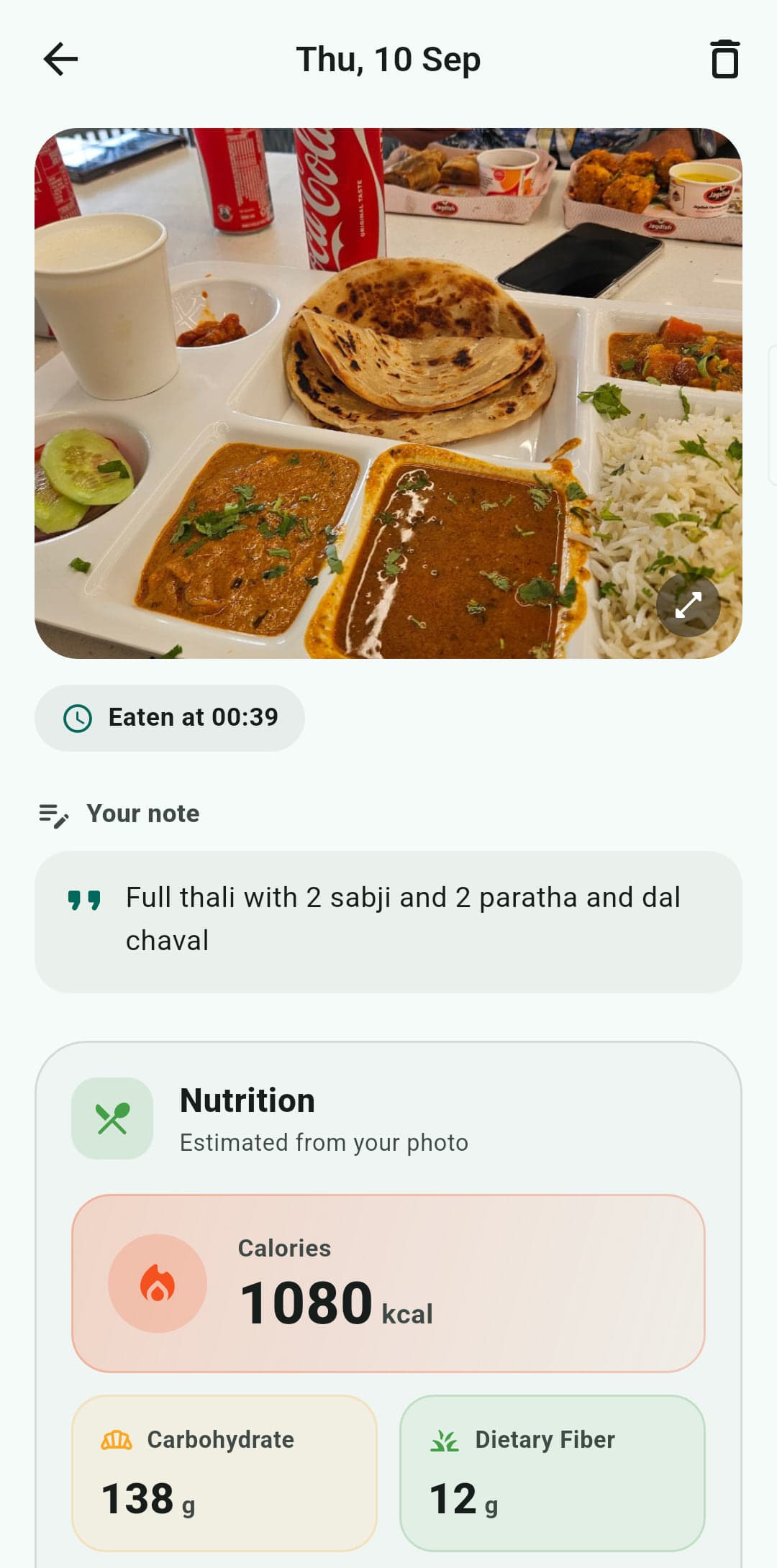 A meal estimated from a photograph: the photo of a thali, the time it was eaten, the patient’s own note reading “Full thali with 2 sabji and 2 paratha and dal chaval”, and a nutrition panel headed “Estimated from your photo” giving 1080 kcal, 138 g of carbohydrate and 12 g of fibre.